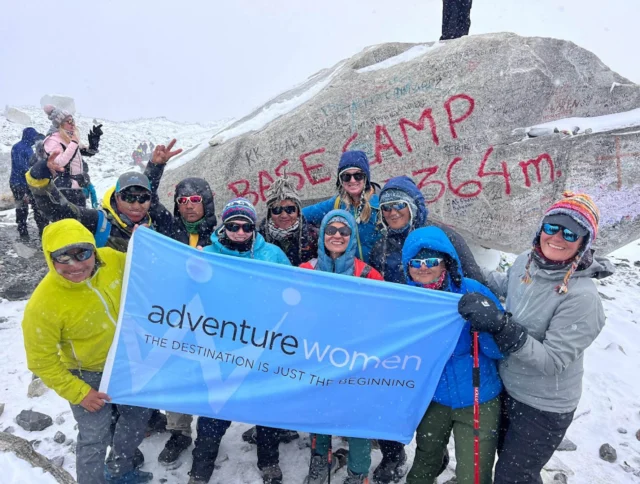 A group of people in winter gear holds a banner reading "adventure women" in front of a large rock with "BASE CAMP 5364m" written on it.