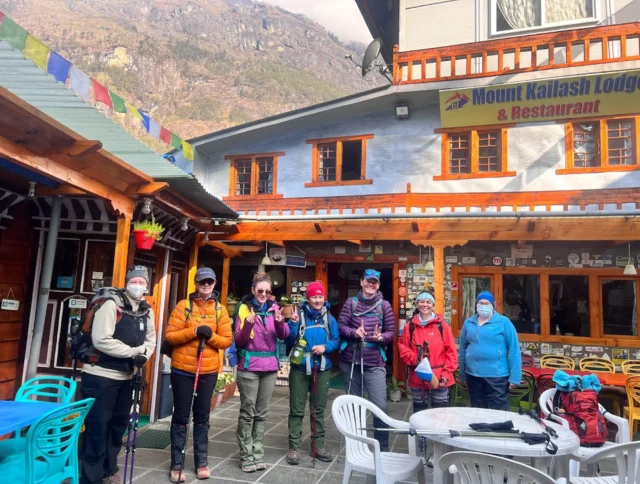 A group of seven hikers with trekking poles stand outside Mount Kailash Lodge & Restaurant in front of a mountainous backdrop.