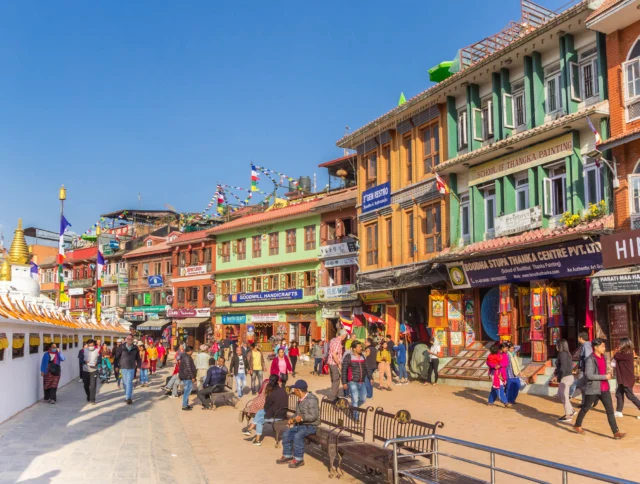 People walk along a colorful street with shops and prayer flags under a clear blue sky.