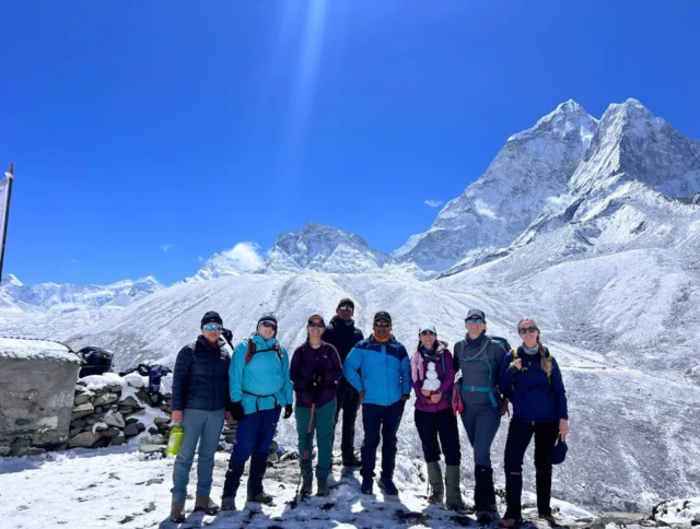 A group of eight hikers stand together on a snowy mountain trail with large snow-capped peaks in the background under a clear blue sky.