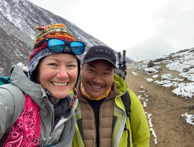 Two people smiling on a snowy mountain trail, dressed in winter gear with colorful hats. The background shows snow-covered hills and a cloudy sky.