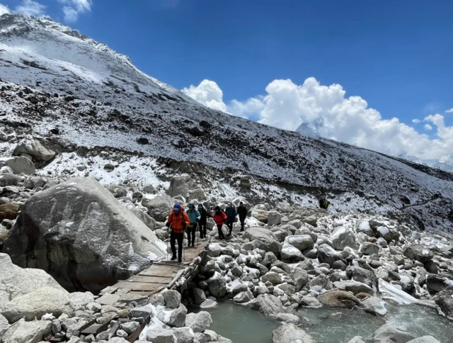 Hikers crossing a narrow wooden bridge over a rocky, snow-covered terrain with a mountainous backdrop under a clear blue sky.