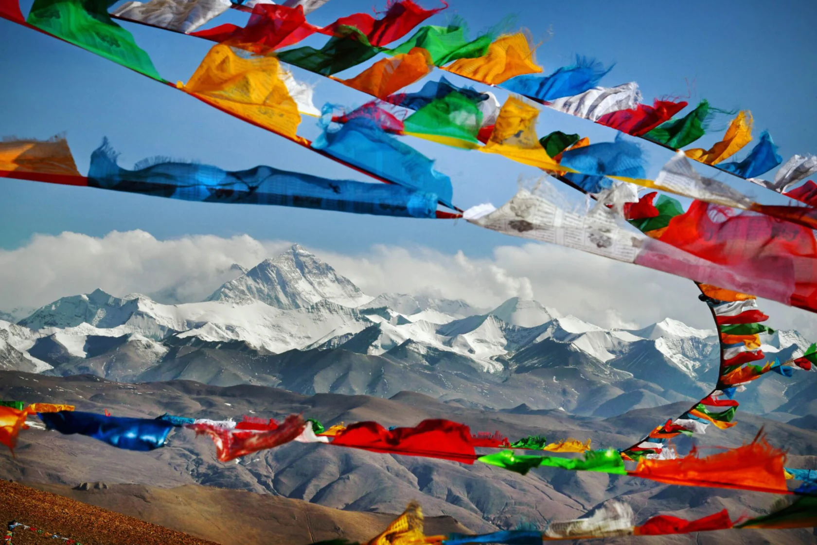 Colorful prayer flags flutter against a backdrop of snow-capped mountains and a clear blue sky.