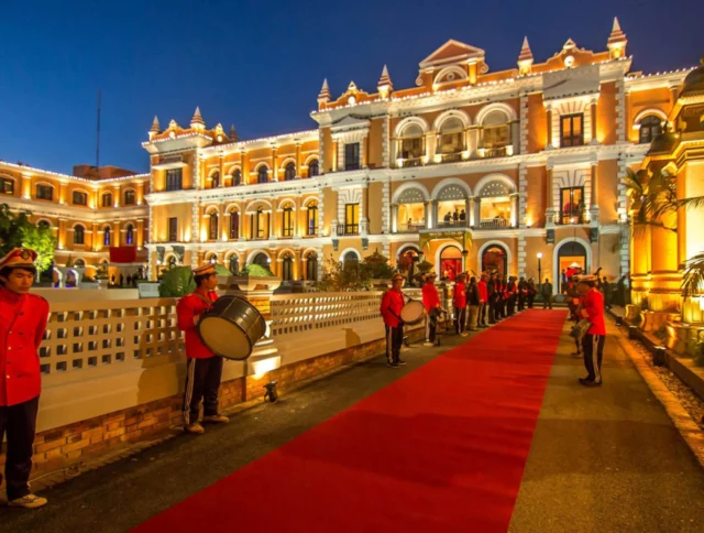 A grand building illuminated at night with a red carpet entrance, lined by uniformed guards holding drums and trumpets.