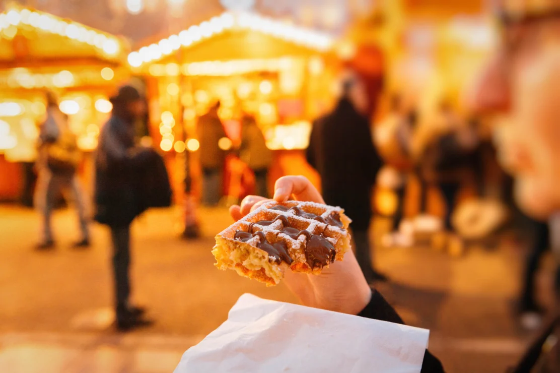 A person holds a partially eaten waffle dusted with sugar. In the background, there are people and brightly lit market stalls.