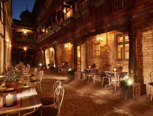 Outdoor courtyard dining area with stone flooring, wrought-iron chairs, tables with tablecloths, and warm lighting. Tables are set with lanterns and flower pots along a rustic wooden balcony.