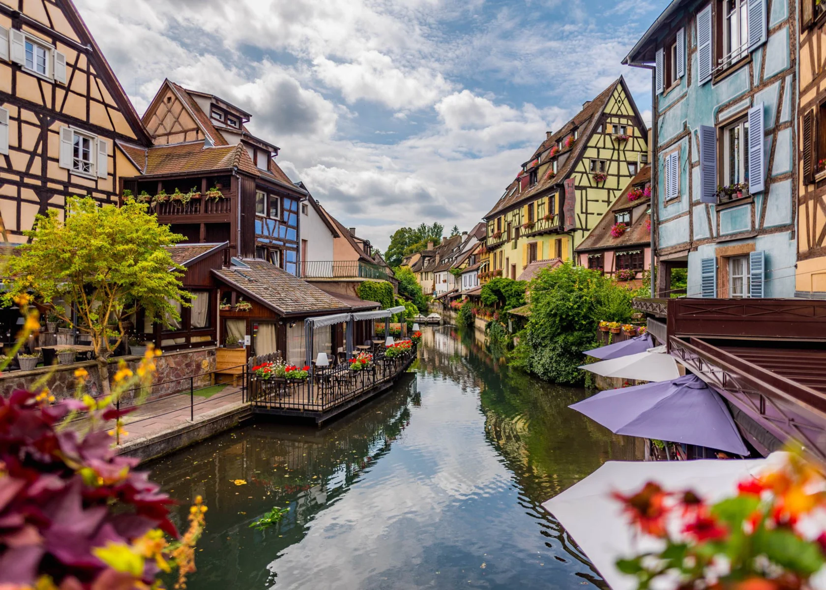 Charming village scene with colorful half-timbered houses lining a narrow canal, surrounded by lush greenery and blooming flowers under a partly cloudy sky.