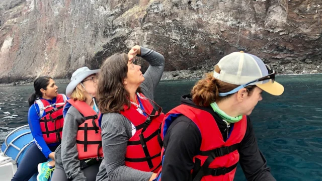 Four people wearing red life jackets sit on a boat near a rocky cliff, with one person pointing towards the sky.