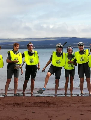 Seven people in wetsuits and yellow vests pose on a beach with snorkeling gear, against a backdrop of water and distant hills.