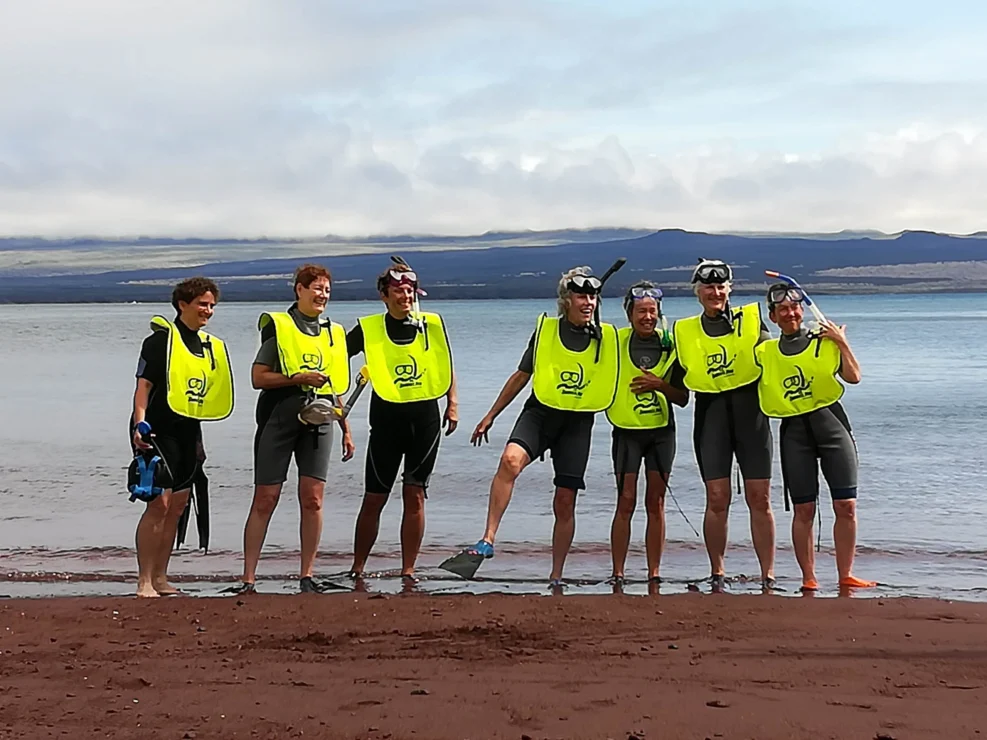 Seven people in wetsuits and yellow vests pose on a beach with snorkeling gear, against a backdrop of water and distant hills.