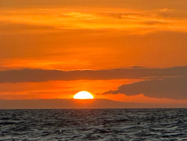 A vibrant orange sunset over the ocean with the sun partially hidden by clouds and a rippling water surface in the foreground.