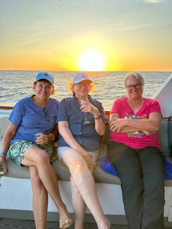 Three women sitting together on a boat, enjoying drinks at sunset with the ocean in the background.