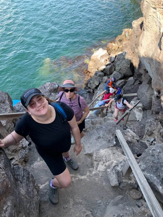 People hiking up a rocky staircase by the ocean, with clear blue water and rocks in the background.