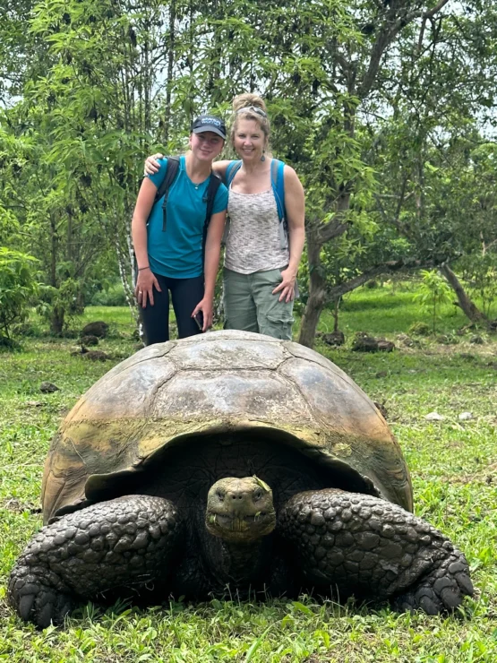 Two people pose behind a large tortoise on grass in a lush, green setting.