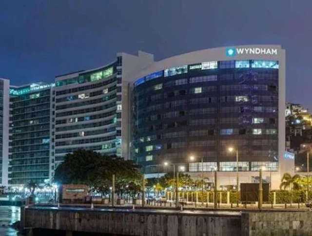 Night view of a modern building with a "Wyndham" logo, illuminated alongside palm trees and streetlights, adjacent to waterfront.