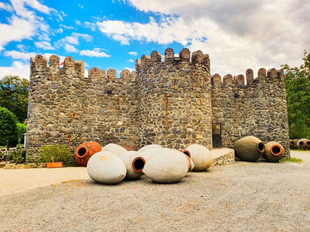 Stone castle with crenellated walls and large, round vessels on the ground, set against a partly cloudy sky.