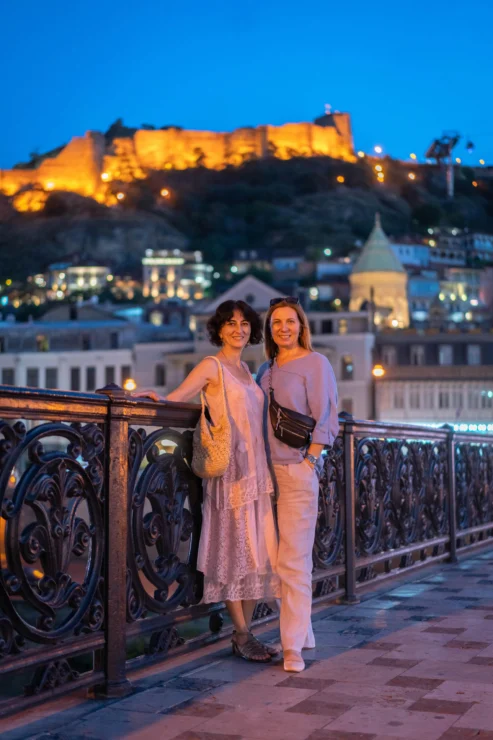 Two women stand on a decorative bridge at dusk, with illuminated historic buildings and a hilltop fortress in the background.