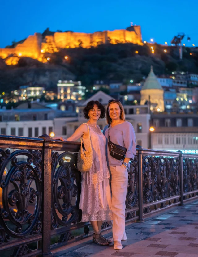 Two women standing on a bridge at night with an illuminated fortress and buildings in the background.