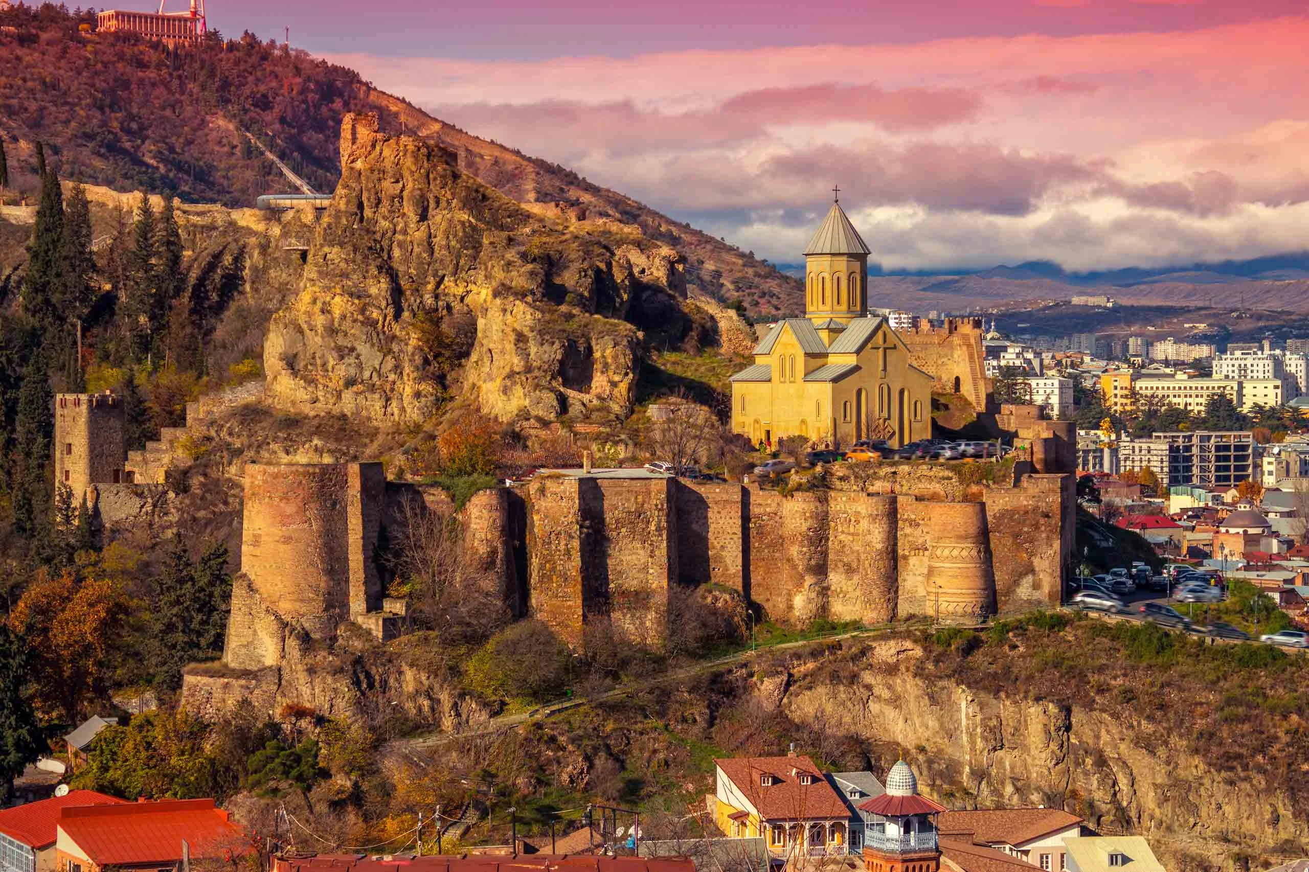 A historic fortress with a church on top of a hill, surrounded by a cityscape under a colorful sky.