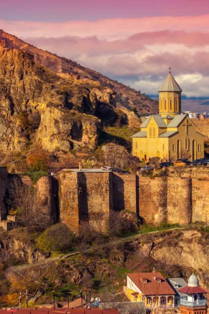 A scenic view of Narikala Fortress in Tbilisi, Georgia, with a church on the hillside, surrounded by city buildings under a vibrant sky.