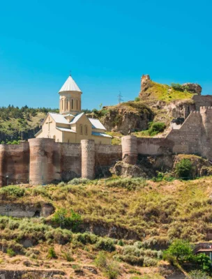 View of an ancient fortress with a church on a hill, surrounded by lush greenery and modern buildings at the base under a clear blue sky.