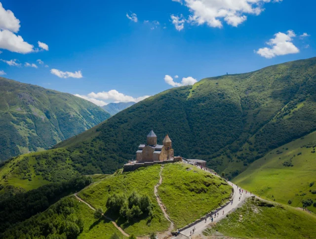 A church sits atop a green hill surrounded by lush, mountainous landscape under a blue sky with scattered clouds.
