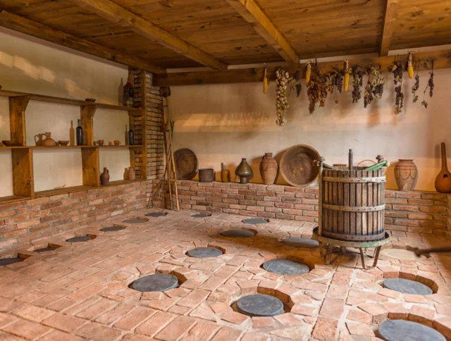 A rustic room with clay pots, jars on shelves, and a wooden wine press. The floor is brick with round holes, and dried herbs hang from the ceiling.