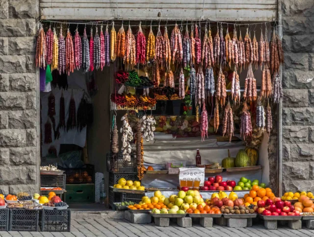 A market stall displays hanging dried sausages and an assortment of fruits including apples, oranges, and lemons on the ground.