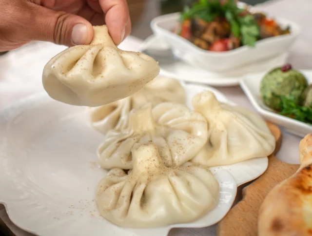 A hand picking up a steamed dumpling from a white plate. More dumplings, a bread roll, and a bowl of greens and vegetables are seen on the table.
