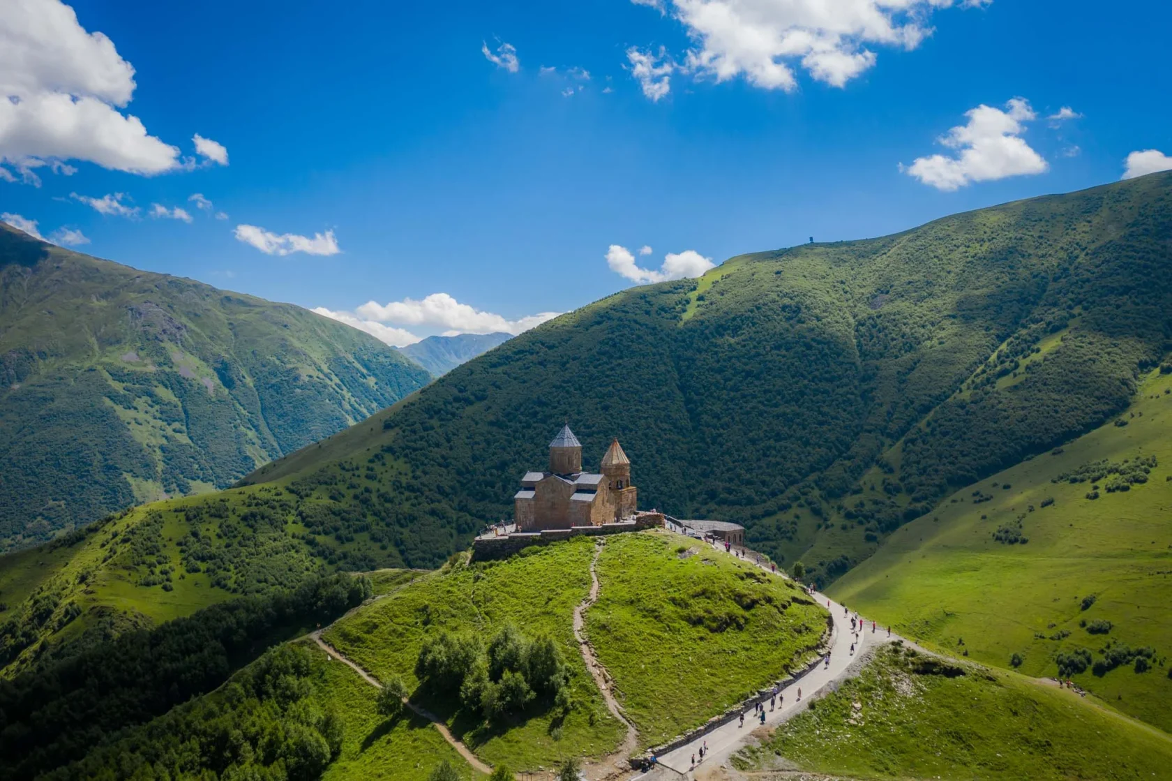 Stone church on a green hilltop surrounded by lush mountains under a bright blue sky with scattered clouds.