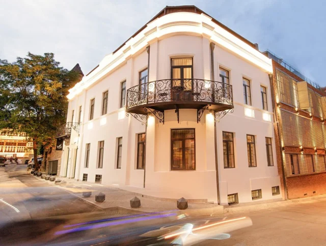 Historic building with a white facade and ornate balcony on a corner at dusk, with light trails from passing vehicles on the street.