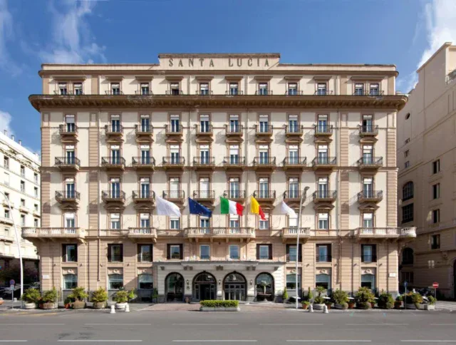 Front view of a multi-story hotel named Santa Lucia, featuring balconies, arched entrances, and several national flags displayed above the main entrance.