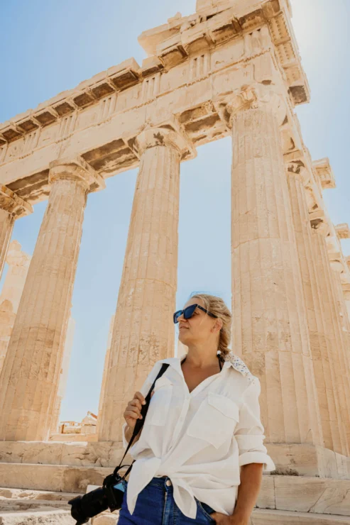 A person in a white shirt and sunglasses stands in front of ancient columns, holding a camera.