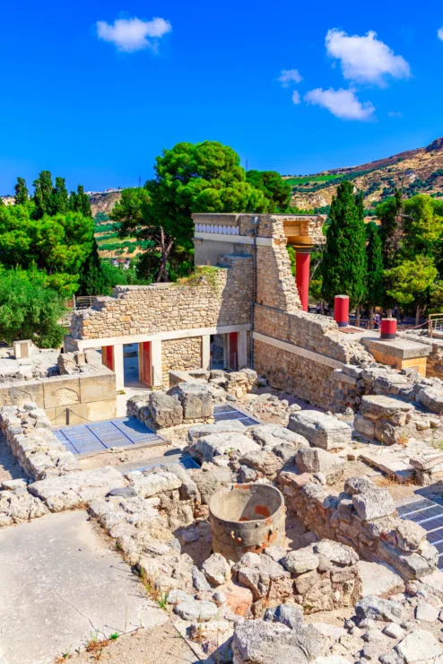 Ancient ruins of Knossos Palace with stone structures, columns, and trees, under a clear blue sky.