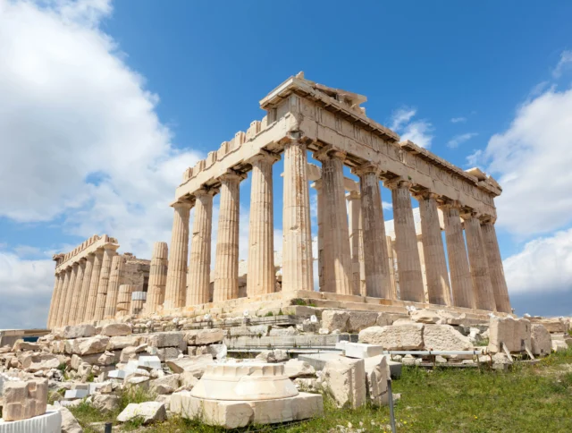 The Parthenon, an ancient temple with Doric columns, sits atop the Acropolis in Athens under a partly cloudy sky. Ruins and scattered stones surround the structure.