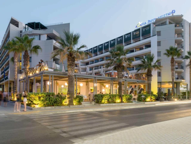 A modern hotel with palm trees stands beside a crosswalk. The building features multiple balconies and an illuminated entrance.