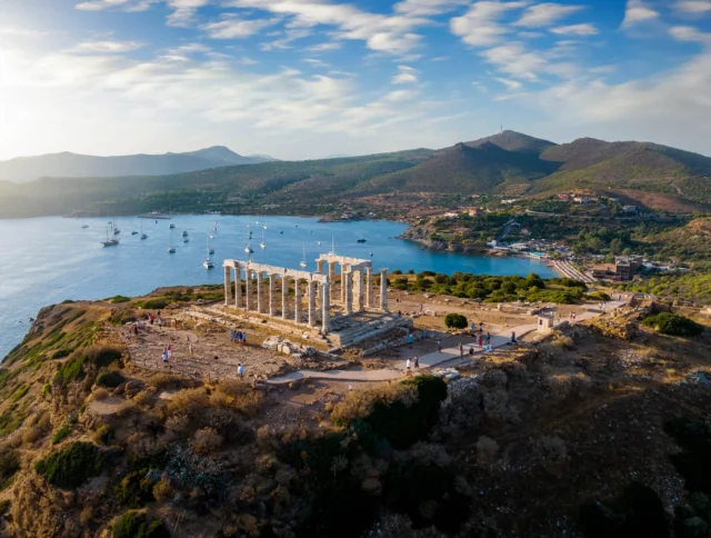 Aerial view of the ancient Temple of Poseidon on a hill overlooking the sea, with surrounding landscape and distant mountains.