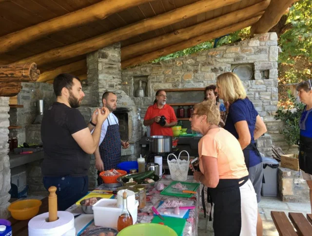 Group of people cooking together in a rustic outdoor kitchen, with ingredients and cookware on the table. Stone walls and wooden roof in the background.