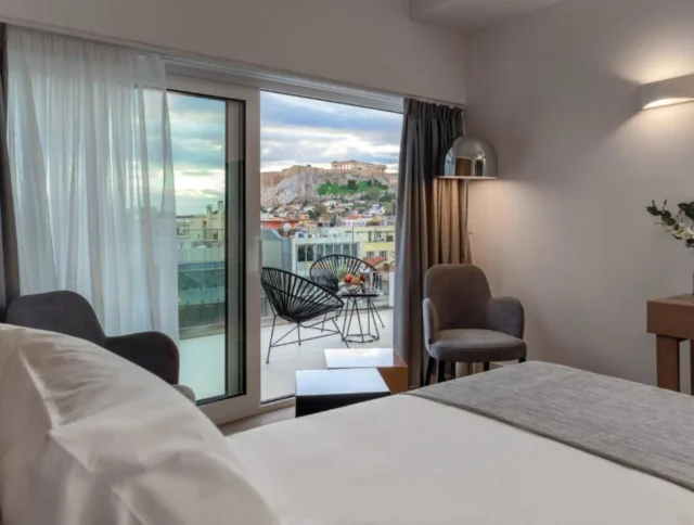 Hotel room with a large bed, two chairs, and a desk. A sliding glass door opens to a balcony with a view of an ancient hilltop structure in the distance.