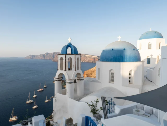 View of Santorini with blue-domed buildings and a bell tower overlooking the sea, with sailboats in the water and a clear sky.