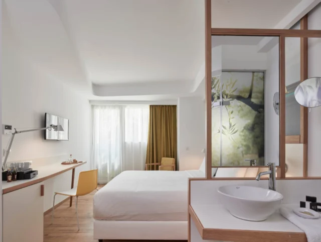 Modern hotel room with a large bed, desk, and chair. There is a sink and mirror near the entrance. Natural light comes through a window with curtains.