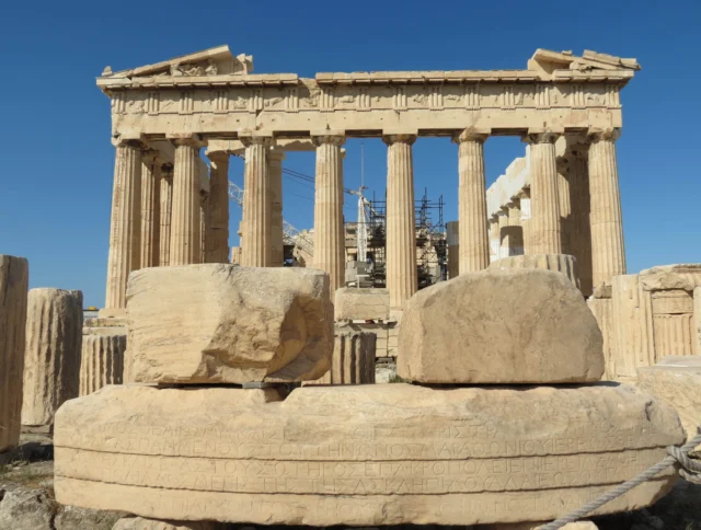 Front view of the Parthenon in Athens, Greece, with ancient stone blocks in the foreground and blue sky in the background.