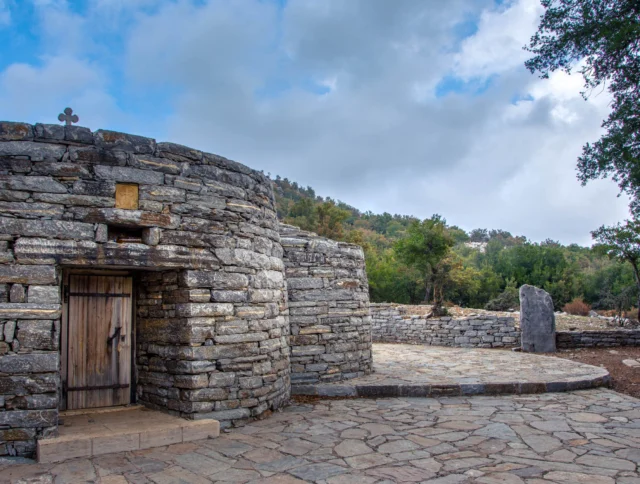 A stone building with a wooden door and a small cross on its roof, surrounded by trees and a stone pathway under a cloudy sky.