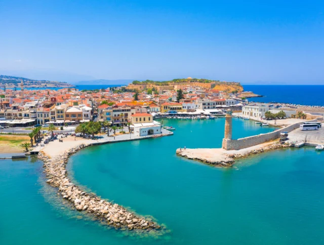 Aerial view of a coastal town with a harbor, surrounded by turquoise water and lined with buildings. A stone pier with a small lighthouse extends into the sea under a clear blue sky.