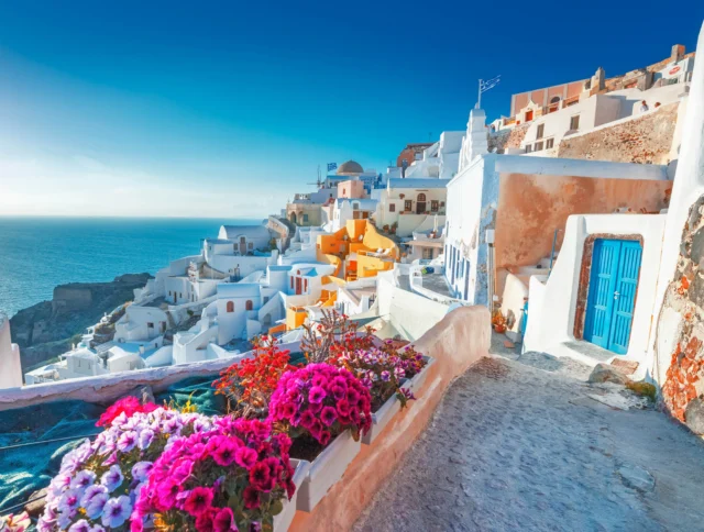 Scenic view of a coastal village with white buildings, a vibrant blue sea, and colorful flowers in the foreground under a clear sky.