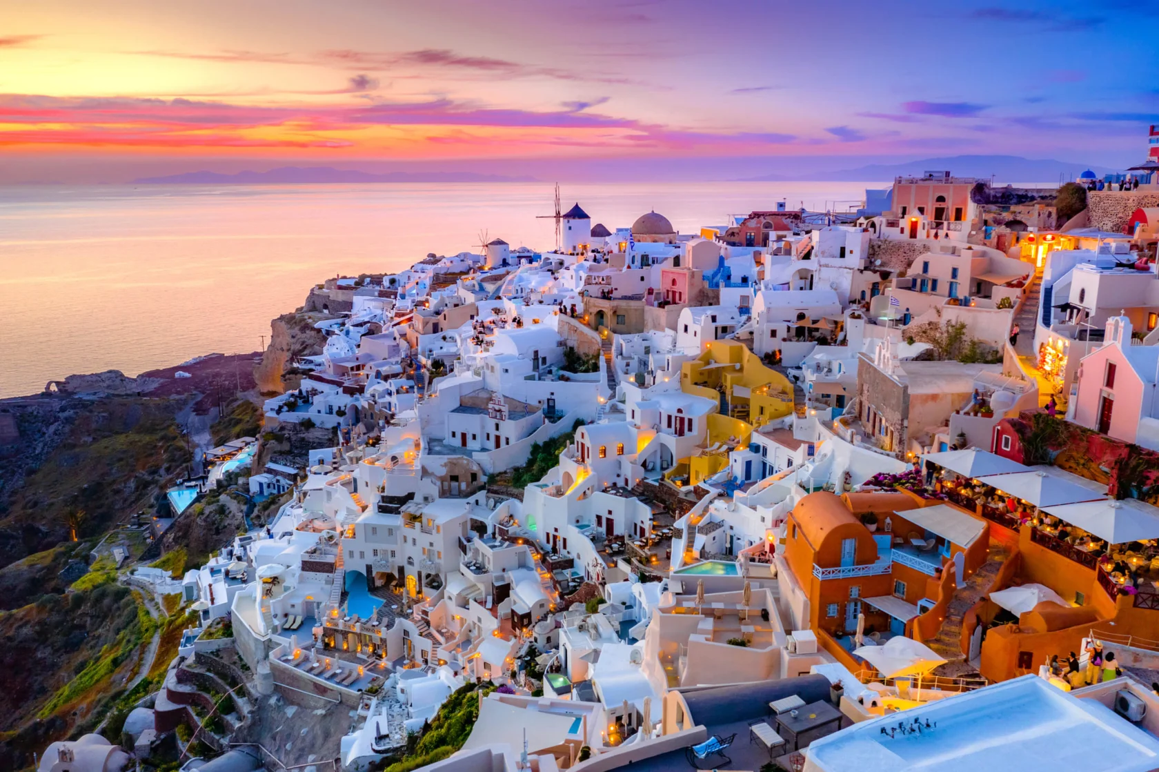 Colorful hillside houses in Santorini, Greece, overlooking the sea at sunset, with a vibrant sky and scattered lights illuminating the buildings.