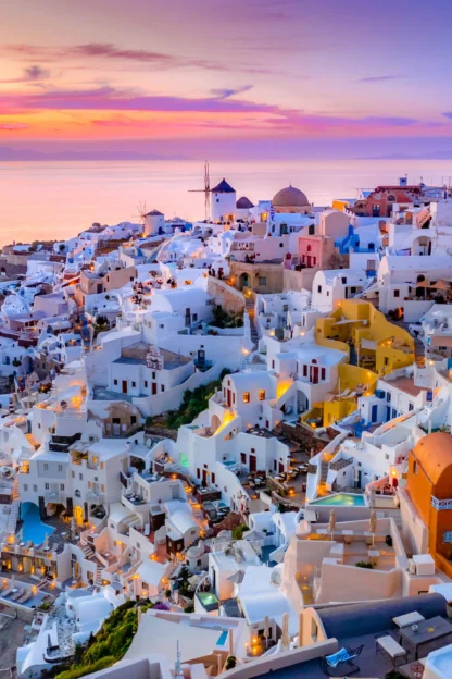 Colorful hillside houses in Santorini, Greece, overlooking the sea at sunset, with a vibrant sky and scattered lights illuminating the buildings.