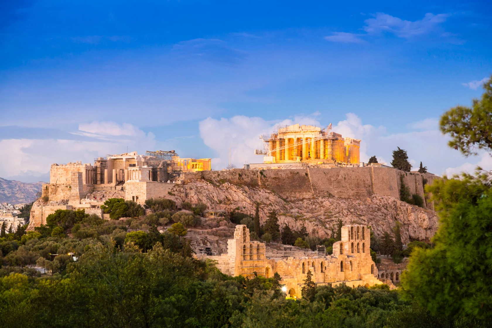 View of the Acropolis in Athens, Greece, featuring the illuminated Parthenon atop a rocky hill with surrounding ruins and greenery under a clear blue sky.