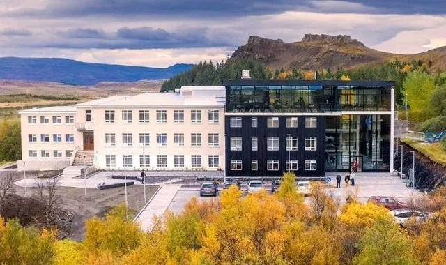 A modern, multi-story building with white and black façades stands among autumn trees and hills under a cloudy sky.