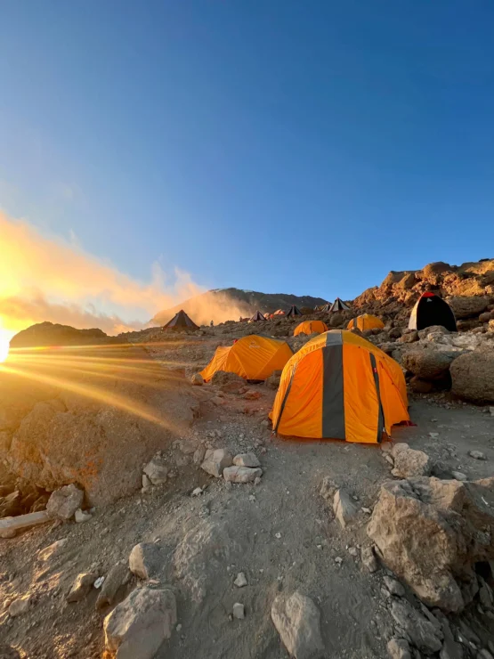 Bright orange tents are set up on a rocky landscape at sunrise, with the sun casting rays over the horizon.